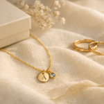 Personalized gold necklace with initial charm and birthstone pendant on ivory linen beside engraved rings and a cream jewelry gift box.