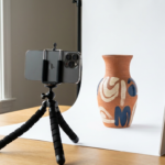 A DIY product photography setup for 2026, showing a smartphone on a tripod, a ceramic vase on a white sweep, and natural window lighting.