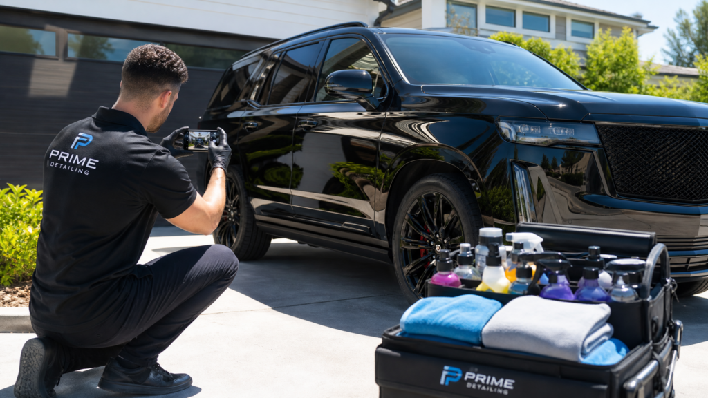 A professional car detailer in a clean uniform using a smartphone to take a photo of a sparkling clean car interior in a sunny driveway.